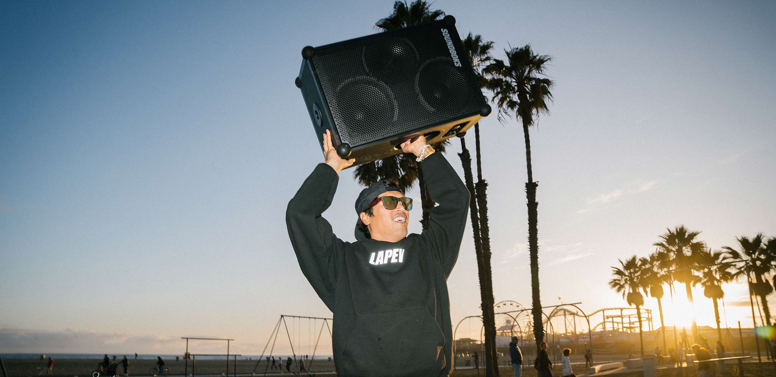 A person wearing sunglasses holds a large SOUNDBOKS 4 speaker overhead. They are standing on a sandy beach with palm trees and people in the background during sunset.