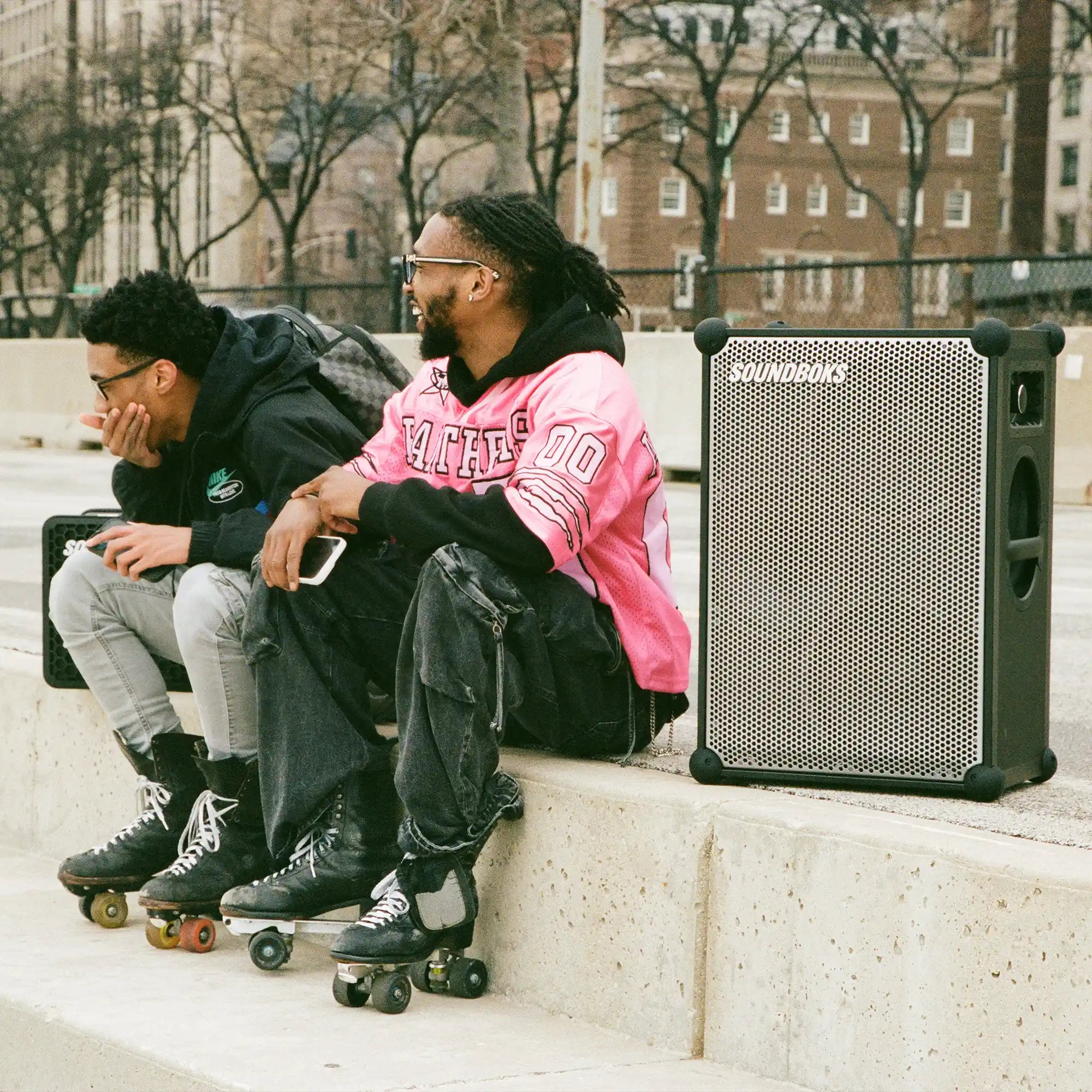 Two people sitting on a ledge with a Metallic grey Soundboks 4 next to them.