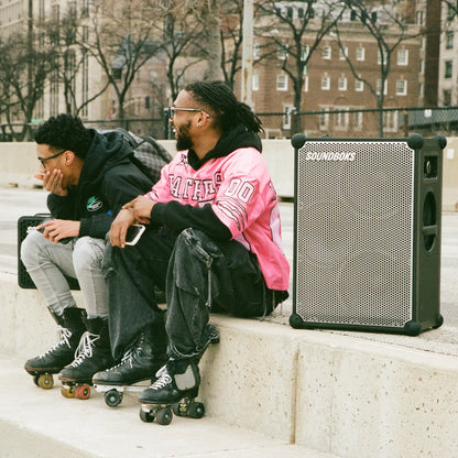 Two people sitting on a ledge with a Metallic grey Soundboks 4 next to them.